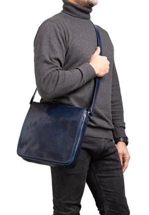 Borsa a tracolla in pelle blu scuro con texture liscia, forma rettangolare e tracolla regolabile, indossata su una spalla.