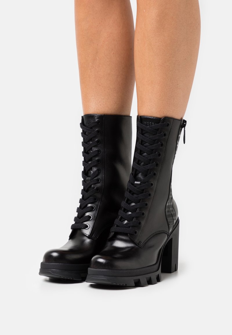 Bottines noires à lacets avec talons hauts épais et fermeture éclair à l'arrière, portées sur des jambes nues contre un fond blanc.