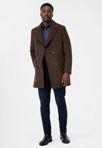 Cappotto in lana marrone con design doppiopetto, ampi rever e bottoni silver, abbinato a una camicia blu navy e pantaloni scuri, completato con stivali neri.