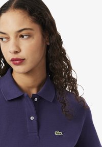 Camisa polo morada con cuello, tapeta de dos botones y un logo de cocodrilo verde bordado en el pecho.