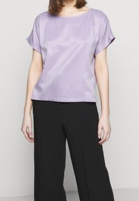 Persona che indossa una blusa in satin lavanda a maniche corte e pantaloni neri larghi, in piedi contro uno sfondo chiaro e uniforme.