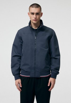 Młody mężczyzna o krótkich włosach, ubrany w granatową kurtkę na zamek z prążkowanymi mankietami i paskowanym dołem, stoi na tle jasnego, jednolitego tła.