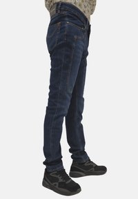 Jeans in denim blu scuro con design slim fit, dettagli cuciti e stile a cinque tasche, abbinati a scarpe sportive nere.
