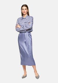 Himmelblaue Bluse mit Muster und Taschen, kombiniert mit einem einfarbigen, himmelblauen Satin-Midirock. Modell trägt graue Flachschuhe mit dekorativem Akzent.