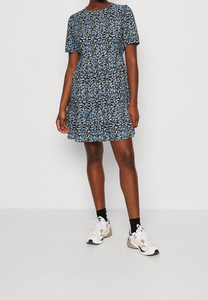 Vrouw draagt een blauw-zwart bloemenjurk met korte mouwen, gecombineerd met witte sneakers en zwarte sokken, staand tegen een eenvoudige witte achtergrond.