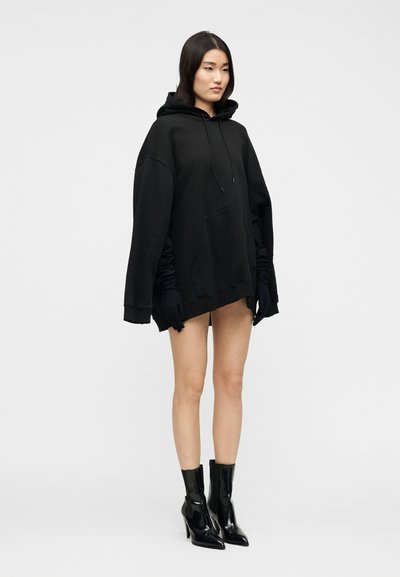 Γυναίκα που φοράει υπερμεγέθη μαύρη φούτερ, μαύρα γάντια και μαύρες ψηλοτάκουνες μποτάκια αστραγάλου, στέκεται σε απλό λευκό φόντο.