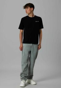 Junger Mann in schwarzem "Tropical Vibe" T-Shirt, hellgrauen Jeans und weißen Sneakers, der vor einem schlichten grauen Hintergrund steht und seitlich schaut.