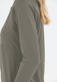 Side view of person wearing long-sleeve olive green jacket with subtle stitching detail and blonde hair visible, on white background.