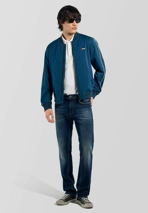 Joven vestido con chaqueta bomber azul oscuro, camisa polo blanca, vaqueros azules, zapatillas negras y gafas de sol, de pie con una mano en el bolsillo.