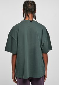 Tricou mare, de culoare verde închis, cu mâneci scurte, textură netedă și decolteu rotund, având o croială lejeră și o terminare dreaptă.