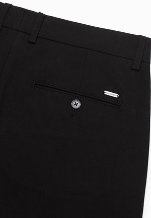 Zwarte broek van textuurstof, met een achterzak met ritssluiting, knoopsluiting en een metalen merketiket op de achterkant.