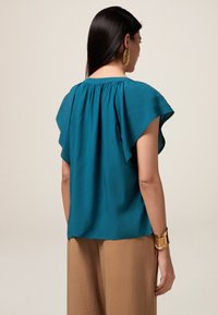 Blusa verde acqua realizzata in tessuto morbido con maniche svolazzanti, scollatura arricciata e vestibilità rilassata. Indossata con pantaloni beige a gamba larga.