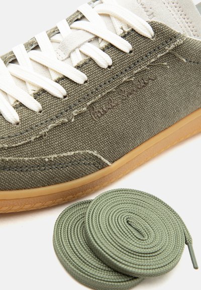 Κοντινό πλάνο σε ελιά καμβά αθλητικό παπούτσι με λευκά κορδόνια και ανοιχτό καφέ σόλα, δίπλα σε δύο τυλιγμένα επιπλέον πράσινα κορδόνια πάνω σε λευκό φόντο.