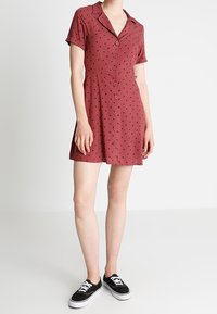 Vestido corto rojo de manga corta con lunares negros, botones en el frente, cuello y cintura ajustada, combinado con zapatillas negras.