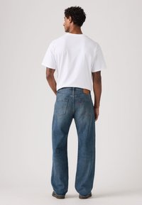 Άσπρο ανδρικό βαμβακερό t-shirt συνδυασμένο με μπλε τζιν παντελόνι, με άνετη εφαρμογή, σχέδιο με πέντε τσέπες και δερμάτινο λογότυπο στο πίσω μέρος.