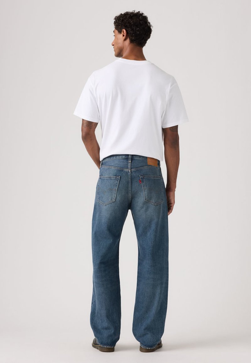 Camiseta de algodón blanca para hombre combinada con jeans de mezclilla azul, con un corte relajado, diseño de cinco bolsillos y parche de cuero con el logo en la parte trasera.