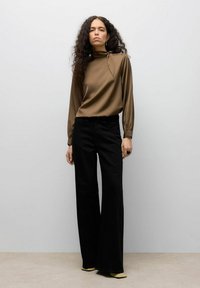 Blouse marron à col montant avec détail noué, pantalon noir à jambes larges et chaussures jaunes à bout pointu, coiffée de boucles lâches et de bijoux minimalistes.