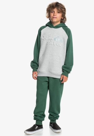 Sudadera verde y gris con un logotipo texturizado, combinada con pantalones de chándal verdes a juego. Zapatillas negras sin cordones completan el atuendo.