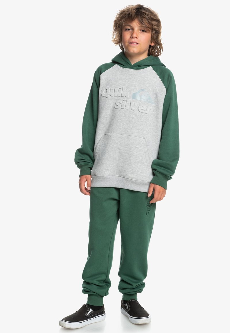 Zelená a šedá mikina s textúrovaným logom, spárovaná so zodpovedajúcimi zelenými teplákmi. Čierne slip-on topánky dokončujú outfit.