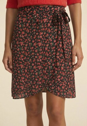 Femme portant une jupe portefeuille noire avec un motif floral rouge nouée à la taille, associée à un pull rouge.