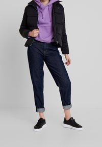 Svart pufferjacka, lila hoodie och mörkblå jeans med uppvikta ben. Svarta sneakers med vita sulor. Avslappnad outfit lämplig för olika tillfällen.