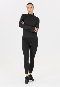 Kvinde står med armene korslagt iført en sort langærmet zip-up top, sorte leggings og sorte sneakers på en hvid baggrund.