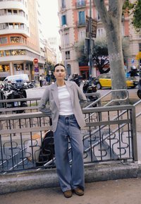 Svetlo sivi blazer čez belo majico, kombiniran s širokimi modrimi kavbojkami. Rjavi čevlji dopolnjujejo opravo. Mesto z zgradbami in vozili.