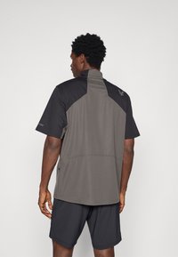 Αθλητικό μπλουζάκι κοντό μανίκι σε γκρι και μαύρο χρώμα. Διαθέτει ψηλό γιακά, αντίθετα πάνελ και διακριτό λογότυπο στο πίσω μέρος.