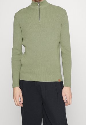 Homme portant un pull zippé côtelé vert olive à col montant et un pantalon noir ample, debout devant un fond clair uni.
