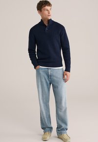 Mörkblå stickad tröja med hög krage och knapprad, tillsammans med ljusblå lösa jeans och beigea slip-on skor.