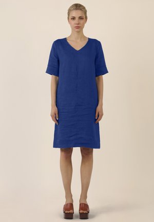 Frau steht in Blickrichtung nach vorn und trägt ein knielanges blaues Kleid mit ellbogenlangen Ärmeln und braune Plateau-Schuhe vor hellem Hintergrund.