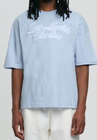 Ljusblå oversized t-shirt i bomull med vit broderad text "Low Lights Studios" över bröstet. Korta ärmar.
