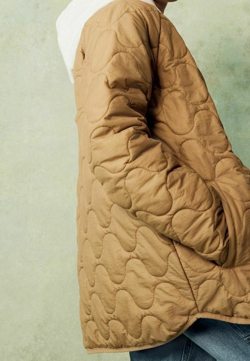 Personne portant une veste matelassée beige avec un motif de couture ondulée, les mains dans les poches, debout contre un mur texturé vert clair.
