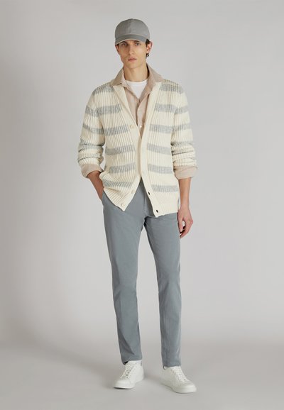 Cardigan lavorato a maglia crema con righe grigie, camicia beige chiaro sotto, pantaloni grigio chiaro e sneakers bianche; un cappello grigio completa il look.