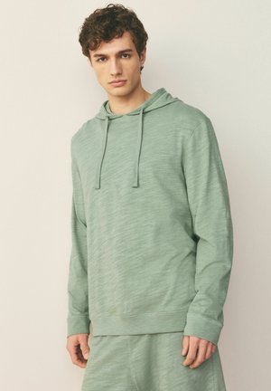 Young man with curly hair wearing a light green hooded sweatshirt and matching pants, standing against a plain beige background.