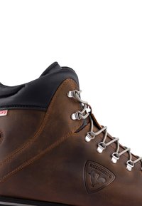 Hnědá kožená turistická bota s černými akcenty, kovovými oky a šedými tkaničkami; má logo vyražené na boku a vysoký design kotníku.