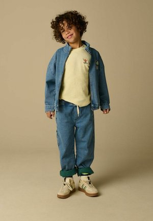 Enfant aux cheveux bouclés debout en souriant, portant un sweat-shirt jaune clair, une veste en denim, un jean retroussé et des baskets beige sur un fond beige.