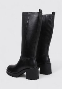 Bottes noires en cuir jusqu'aux genoux avec un talon épais, surface texturée et détails de couture verticales. Équipées de languettes pour un enfilage facile.