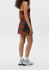 Robe noire à motif floral avec de grandes fleurs rouges et des feuilles vertes, avec des bretelles fines et un col volé. Portée avec des baskets blanches.