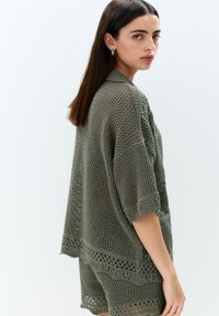Olivgrön virkade topp med korta ärmar, öppen vävdesign, vågig nederkant och intrikata mönsterdetaljer.