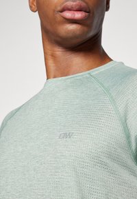Camiseta atlética de color verde claro con tejido texturizado, cuello redondo y logotipo sutil. Presenta un patrón de malla transpirable y mangas raglán.