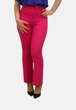 Donna che indossa pantaloni aderenti rosa acceso e tacchi neri appuntiti, con una mano sui fianchi e l'altra appoggiata sulla coscia.