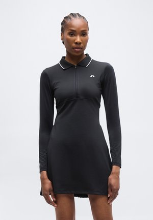 Femme aux cheveux tressés portant une robe polo noire à manches longues avec un petit logo blanc sur la poitrine, sur un fond clair uni.
