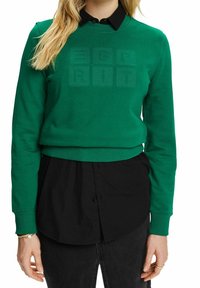Sudadera verde con mangas largas, puños acanalados y un corte corto. Presenta un sutil patrón de texto en el centro. Llevada sobre una camiseta negra.