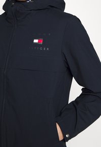 Giacca blu navy con chiusura a zip, cappuccio e tasche frontali. Presenta un logo stampato e un dettaglio a blocchi di colore sul petto. Tessuto morbido.