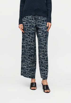 Nő, aki széles szárú tengerészkék nadrágot visel fehér absztrakt vonal mintákkal, egy texturált tengerészkék pulóvert, és fekete, nyitott orrú magassarkú papucsot.