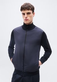 Νέος άνδρας με κοντά μαλλιά, φοράει σκούρο μπλε μπουφάν με φερμουάρ και μακριά μανίκια, στέκεται με τα χέρια στις τσέπες μπροστά σε απλό φόντο.