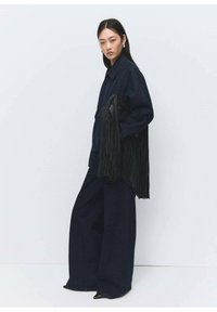 Nainen pukeutuneena löysään tummansiniseen farkkutakkiin, jossa on pitkät mustat hapsut hihoissa, ja leveälahkeisiin, samaa materiaalia oleviin housuihin, seisoo sivuttain valkoista seinää vasten.