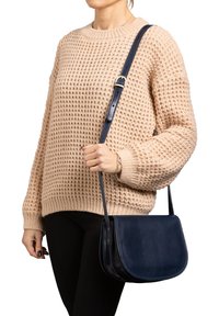 Person in einem beigen Strickpullover und schwarzen Hosen, der eine marineblaue Leder-Umhängetasche mit verstellbarem Tragegurt hält.