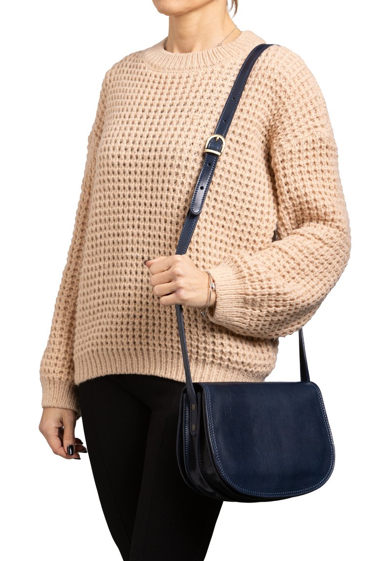 Person in einem beigen Strickpullover und schwarzen Hosen, der eine marineblaue Leder-Umhängetasche mit verstellbarem Tragegurt hält.